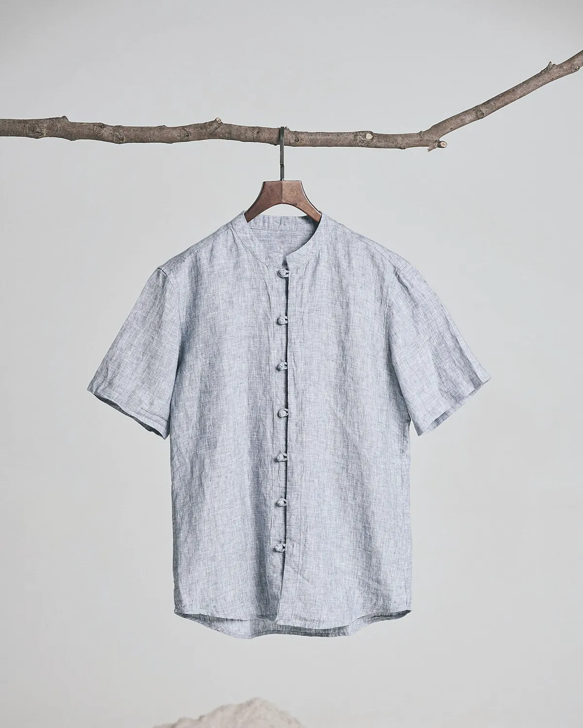 A short sleeve chambray linen shirt with a band collar and a row of fabric knot buttons on a hanger hung from a branch