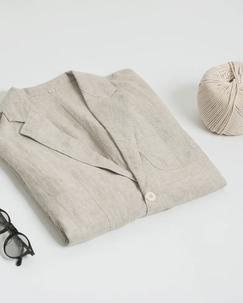 A natural linen overshirt with a notch collar folded on a white table, beside a ball of twine and a pair of glasses