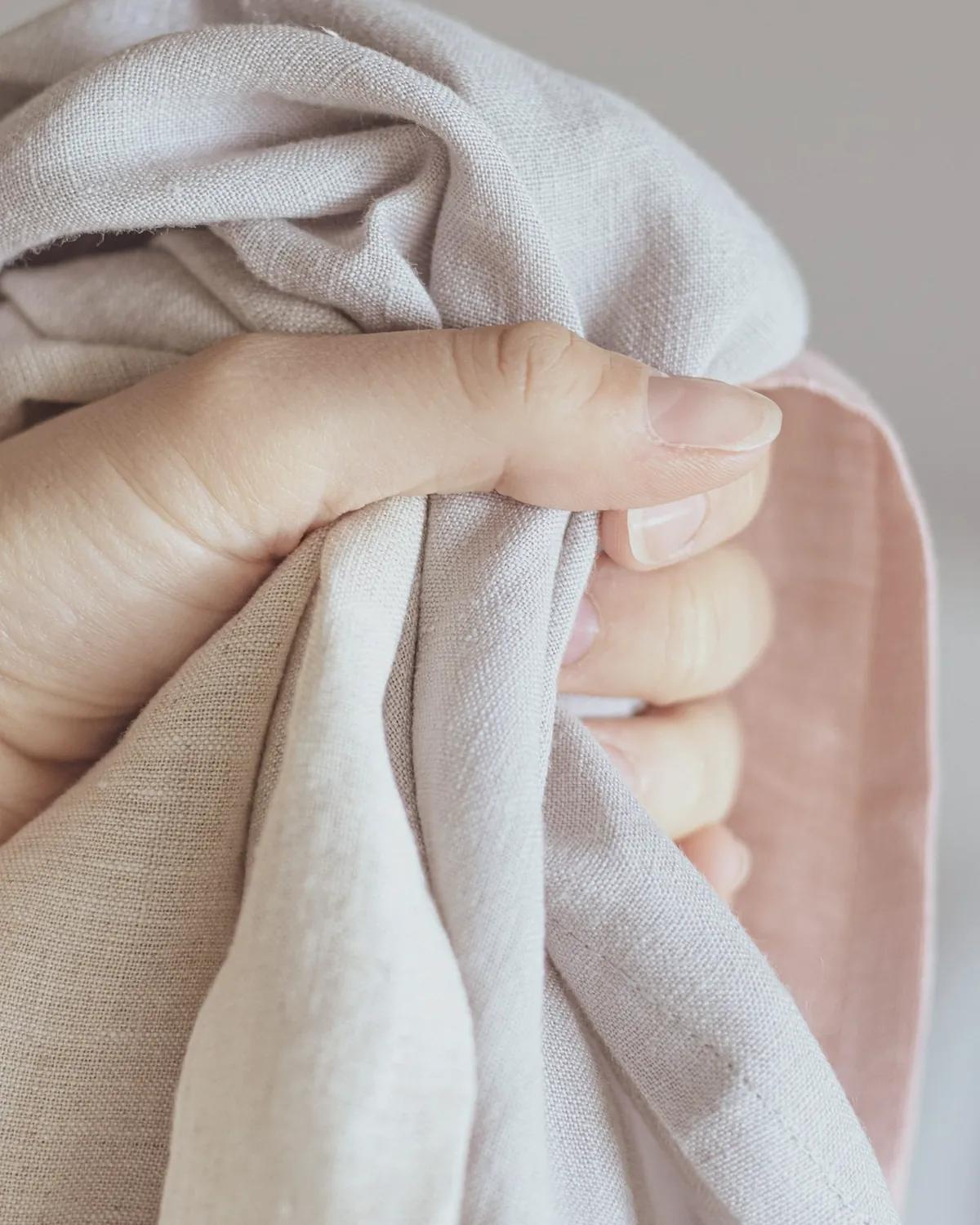 A hand gathers a light undyed linen scarf with a blush pink border