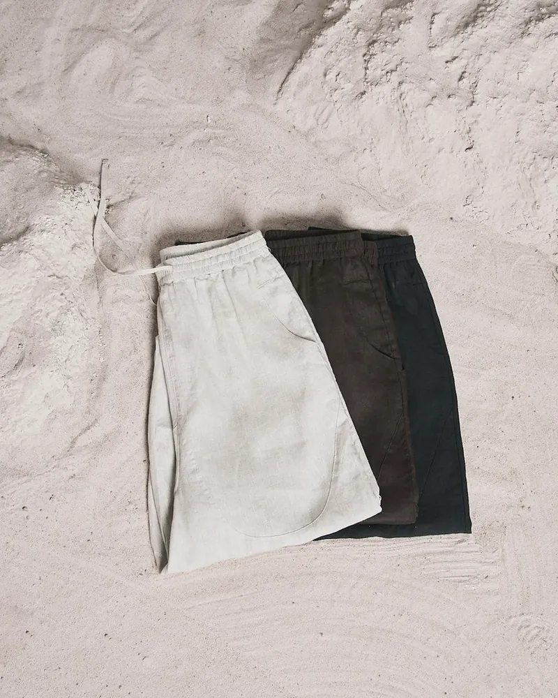 Three pairs of linen drawstring shorts in natural, walnut and black laid out on pale sand