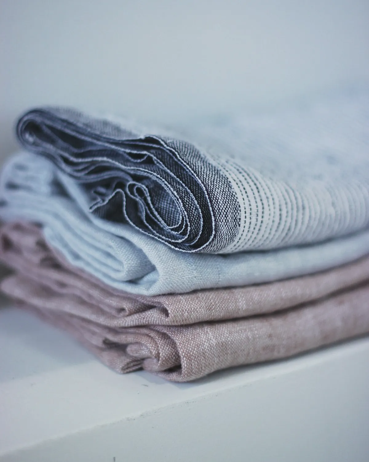 Folded linen tea towels, one with a blue stripe and one in plain mauve, stacked on a shelf