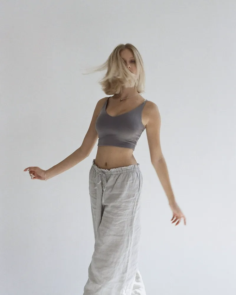 A woman in wide pale grey linen trousers with a drawstring waist turns in a white studio