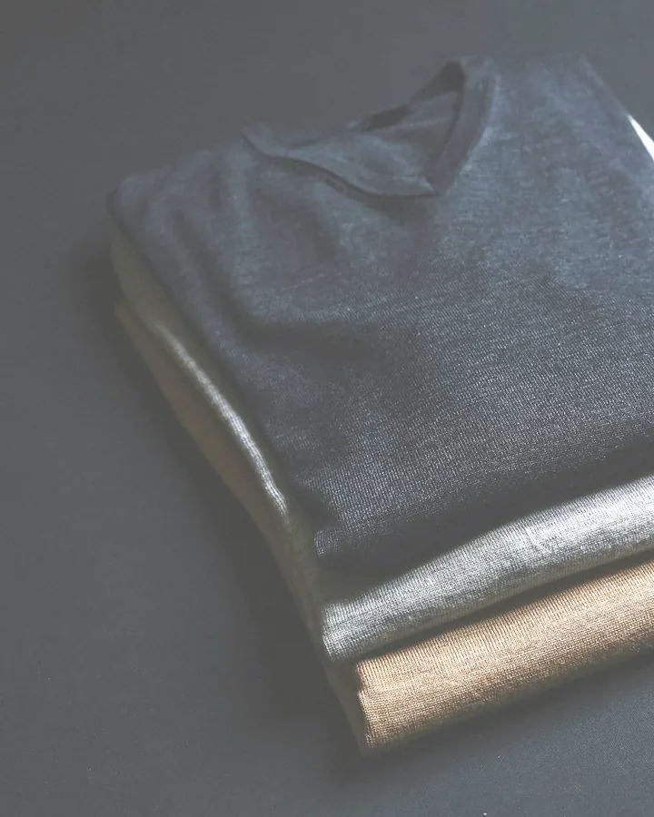Three folded fine knit jumpers in charcoal, grey and camel on a dark table