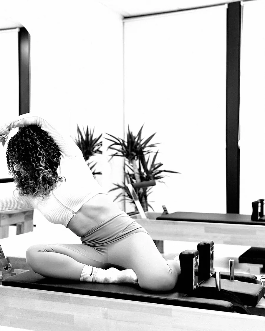 A woman with curly hair stretching on a reformer, seen from behind
