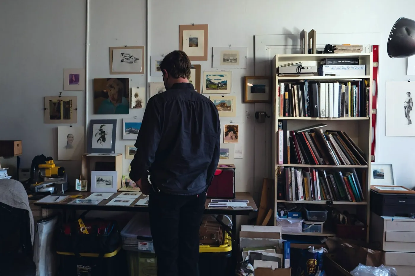A man seen from behind at a work table in a studio full of small framed pictures and books