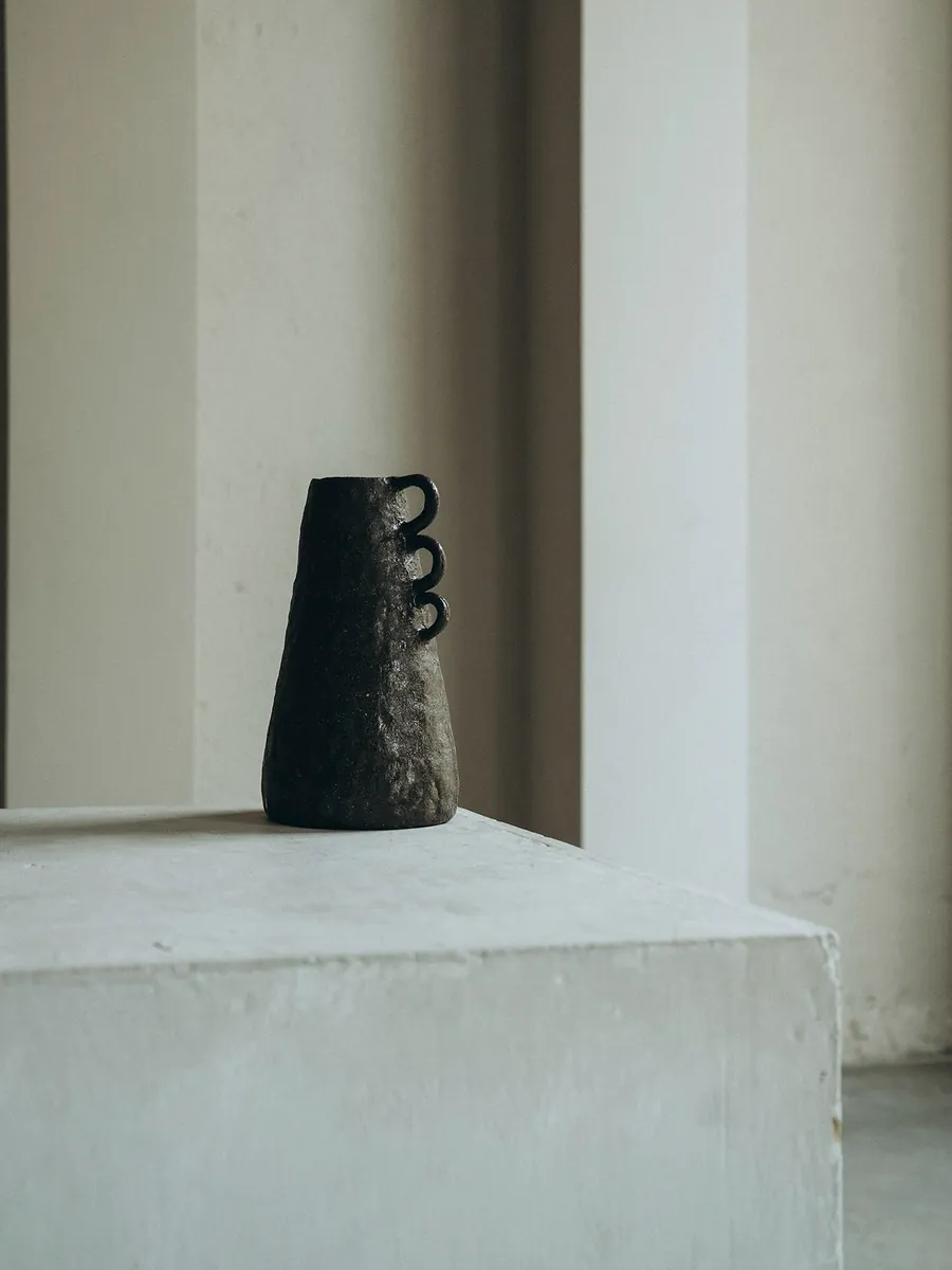 A black textured stoneware jar with three loop handles, standing on a white plinth