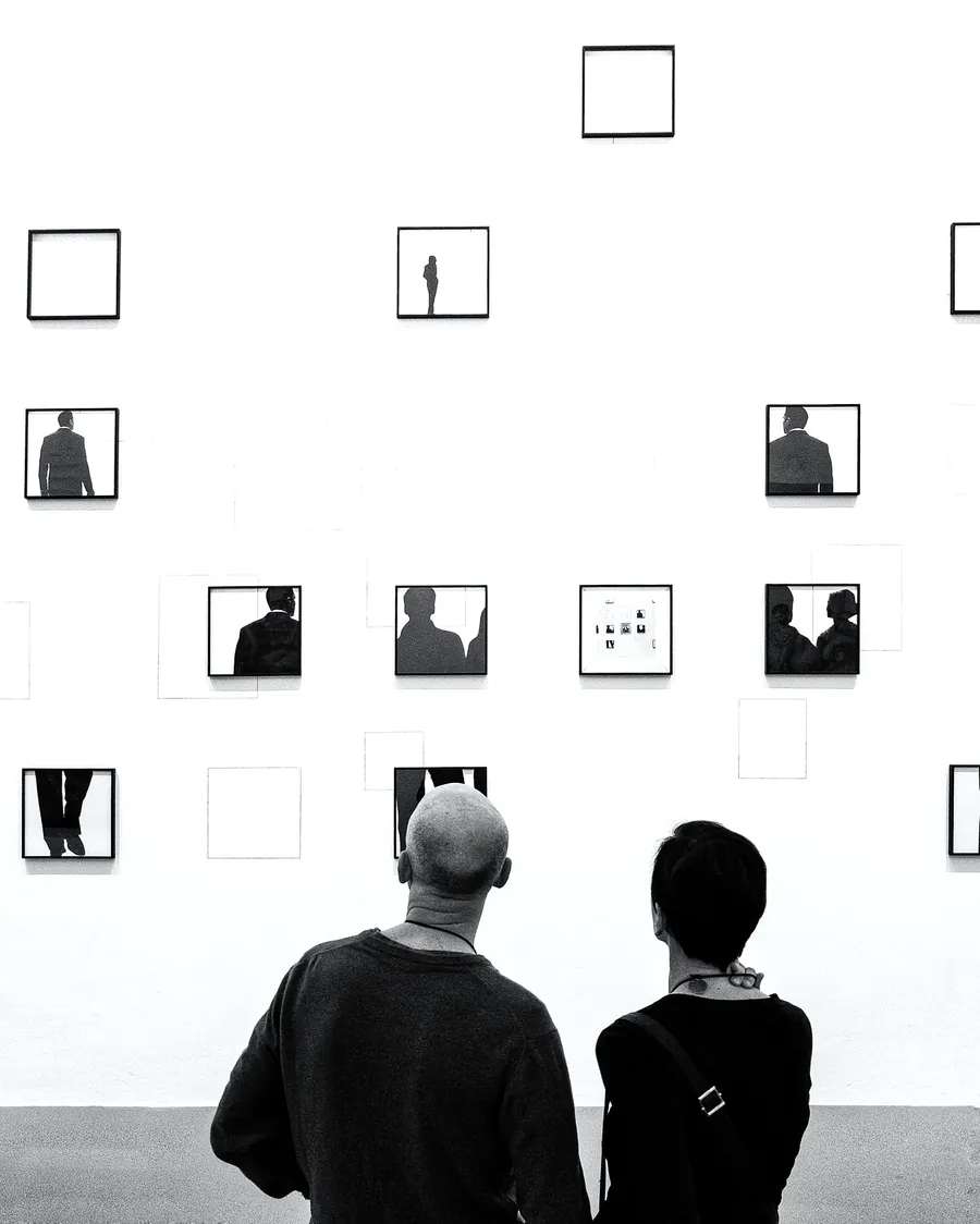 Two visitors seen from behind in front of a wall of small framed works, in black and white