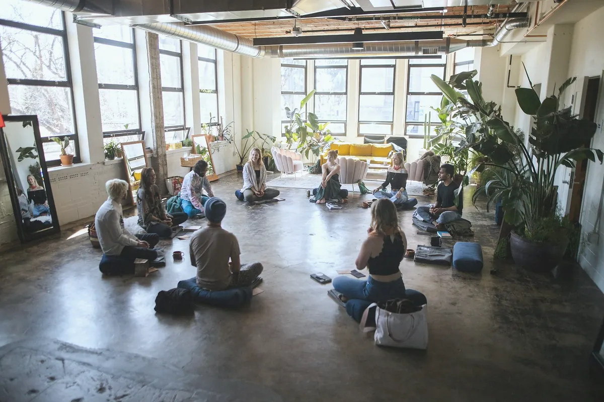 A small class sitting on mats in a wide hall with tall windows and large plants