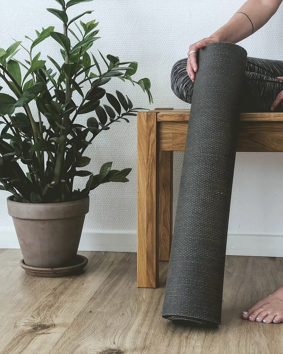 Someone sitting on a wooden bench holding a rolled grey mat, next to a potted plant