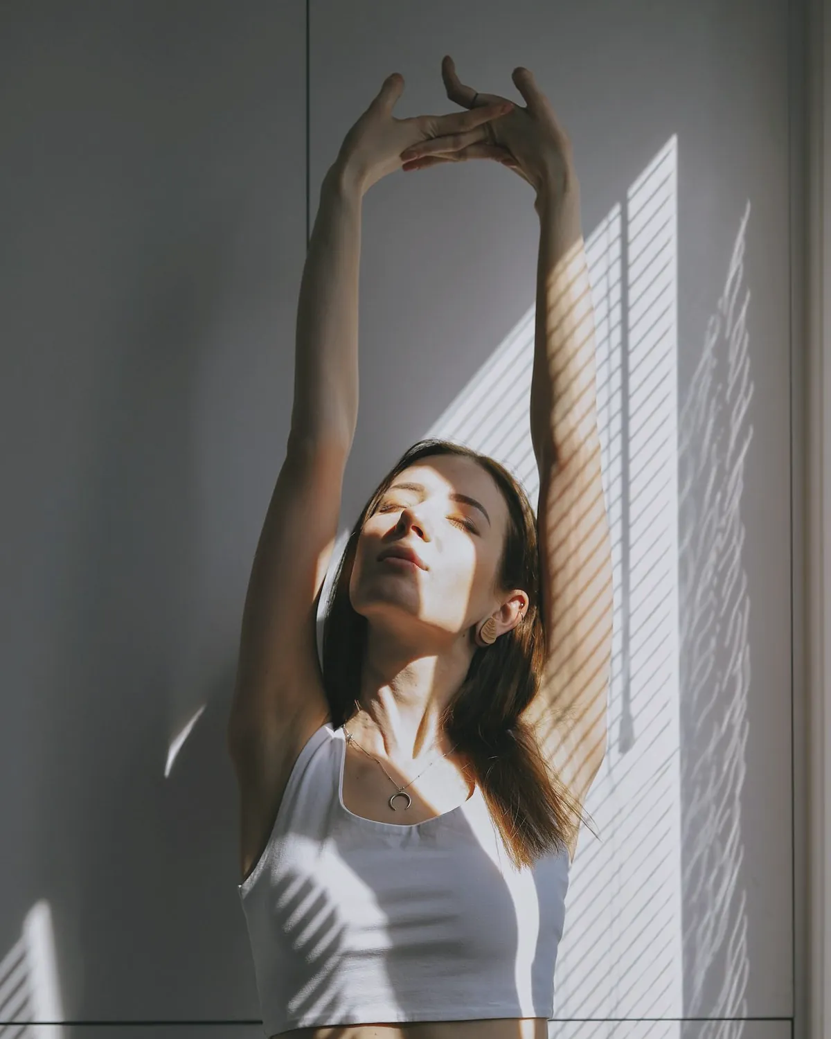 A woman stretching both arms overhead against a wall, eyes closed, with window light across her