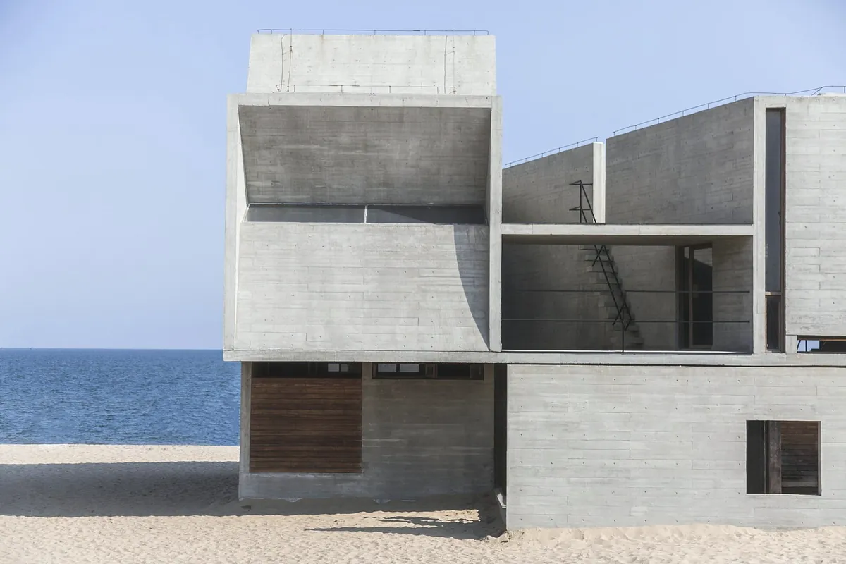 A board-marked concrete house with two cantilevered upper rooms and timber shutters, on a sandy plot by the sea