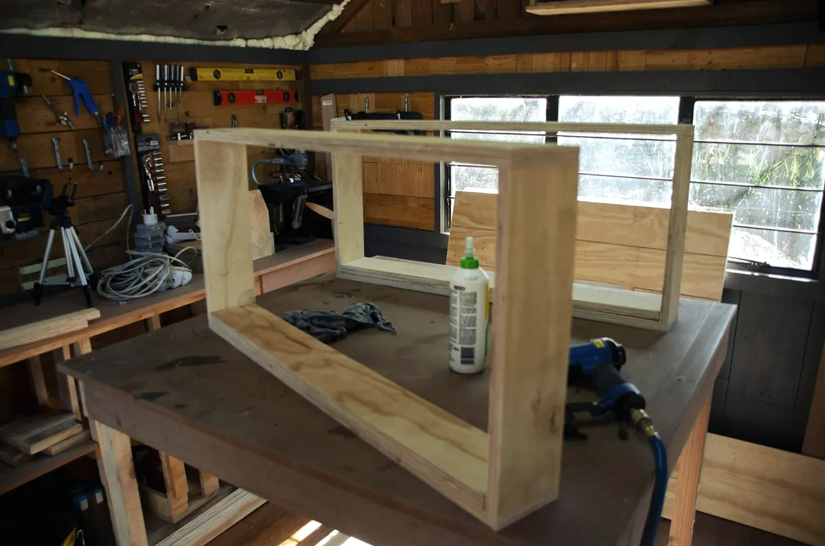 A plywood cabinet frame on a workbench with a glue bottle and an air nailer, in a workshop