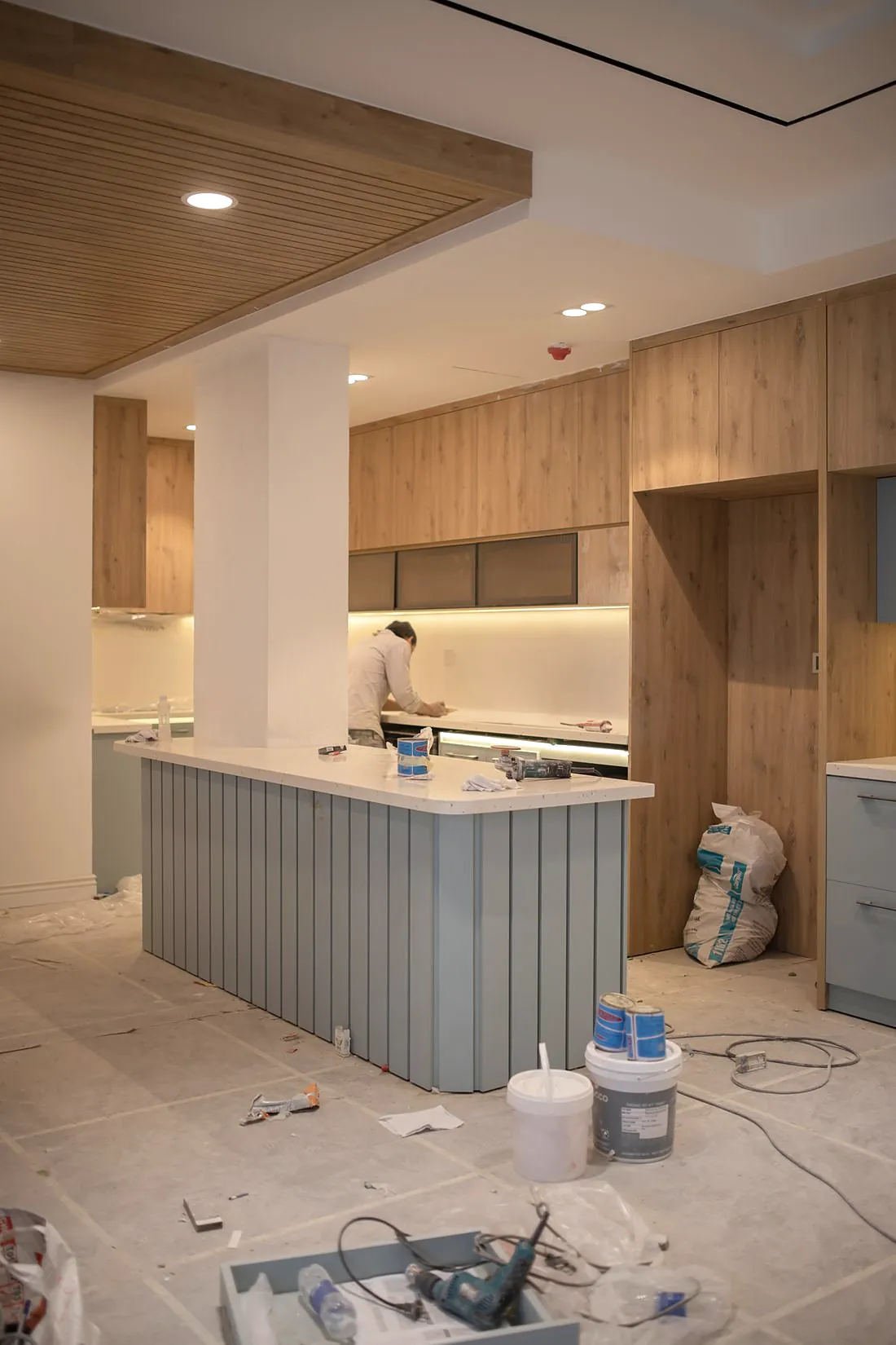 A kitchen being fitted, with timber wall cabinets, a pale blue island and paint tins on the floor