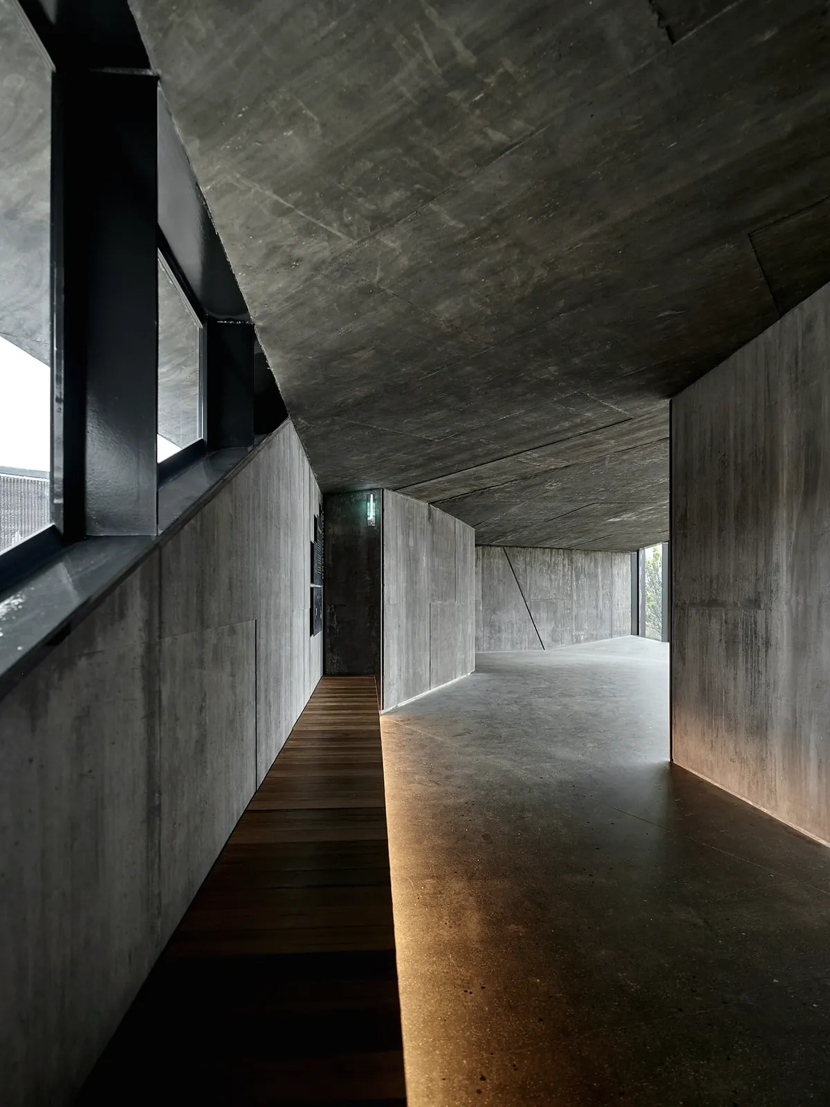 A concrete hallway with a dark timber floor strip, a low concrete ceiling and a lit window at the far end