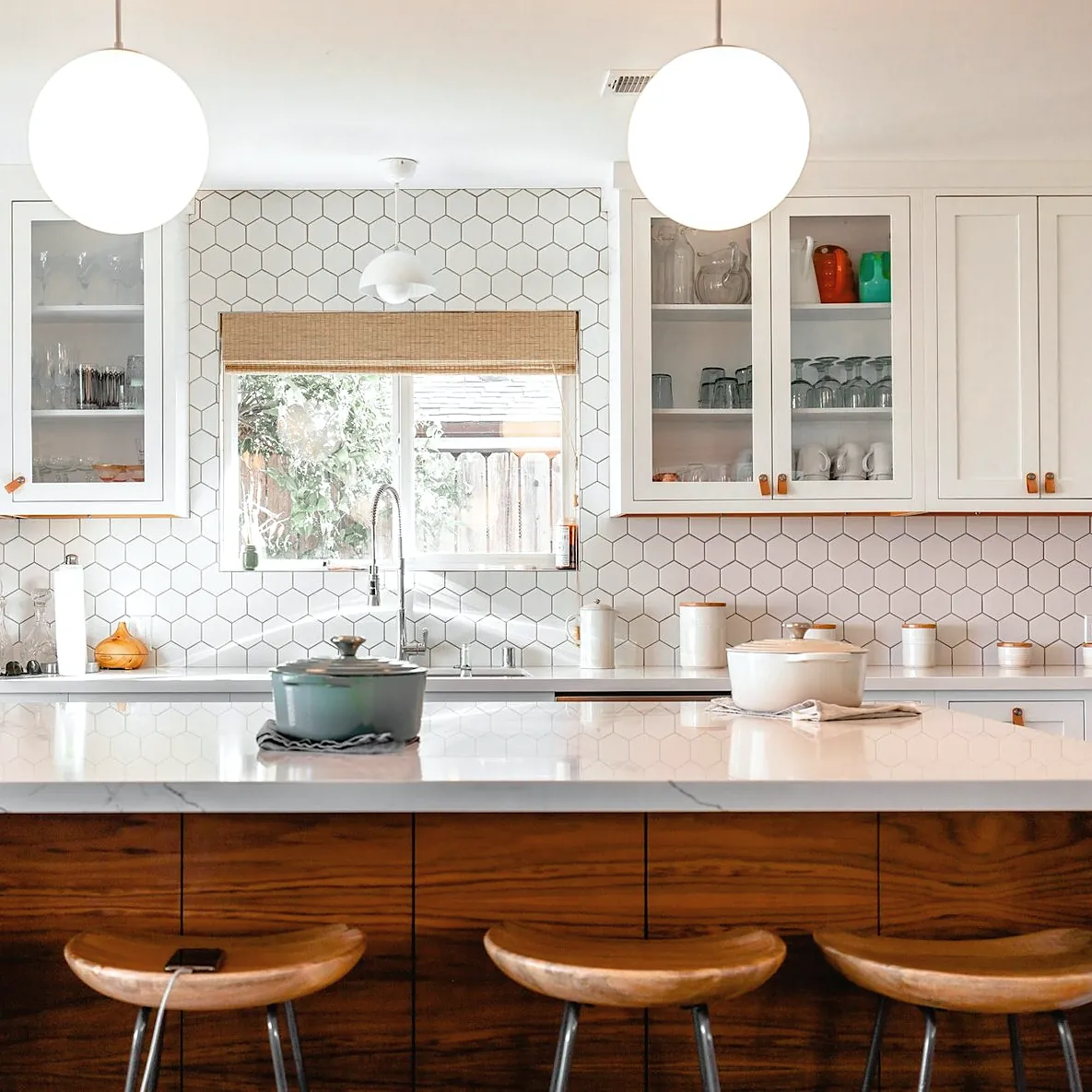A kitchen with a long island with a white stone top and a dark teak front, four wooden stools, glass-front cabinets and a window over the sink