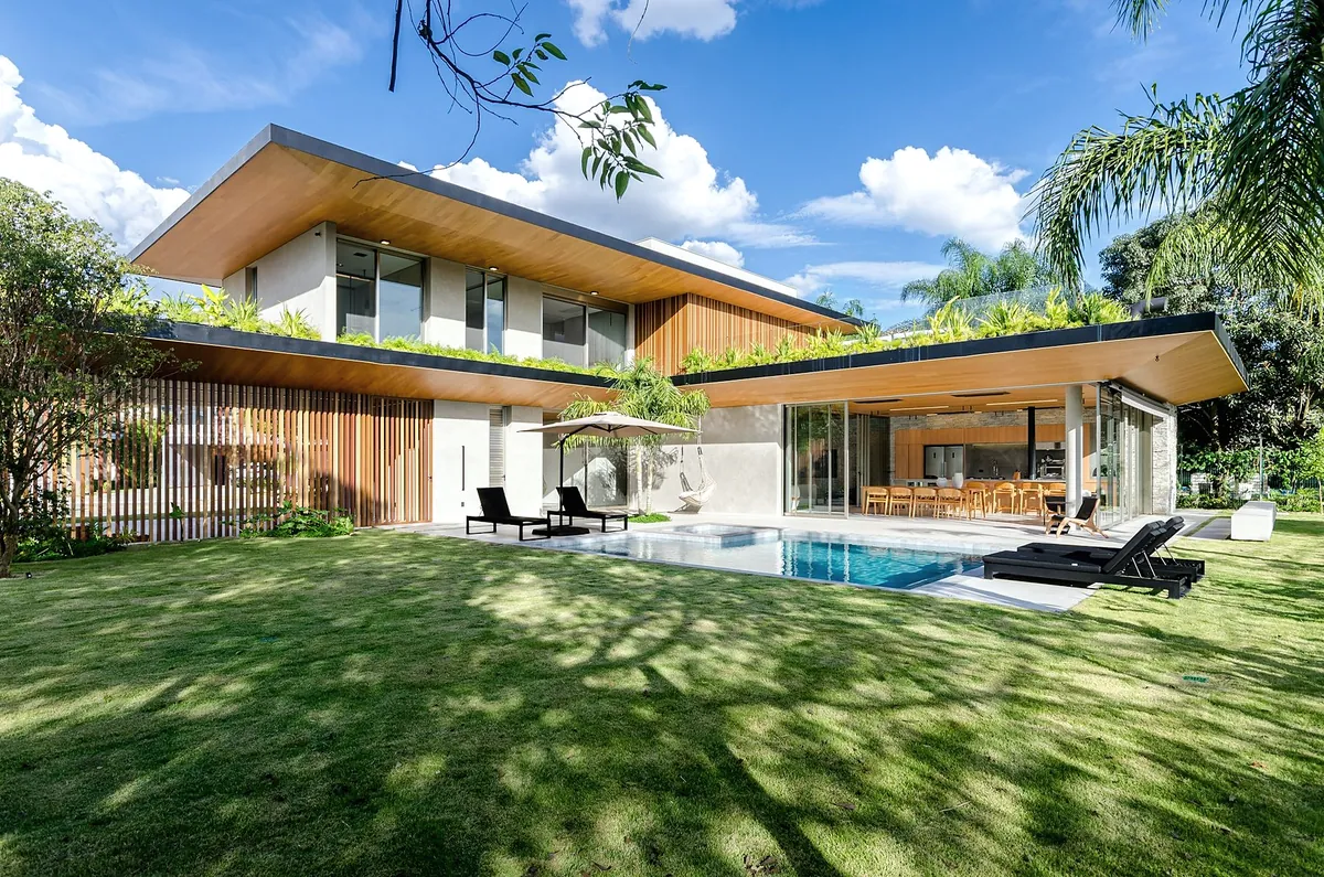 A two-wing house with a long, low timber-lined roof, glass walls, a pool and a lawn with palm trees