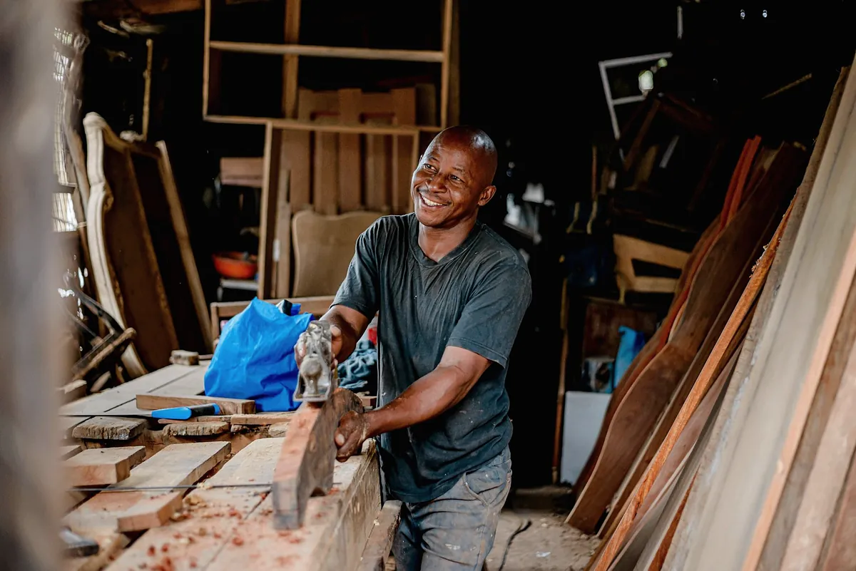 A carpenter planing a thick hardwood board by hand at a bench in a workshop full of timber