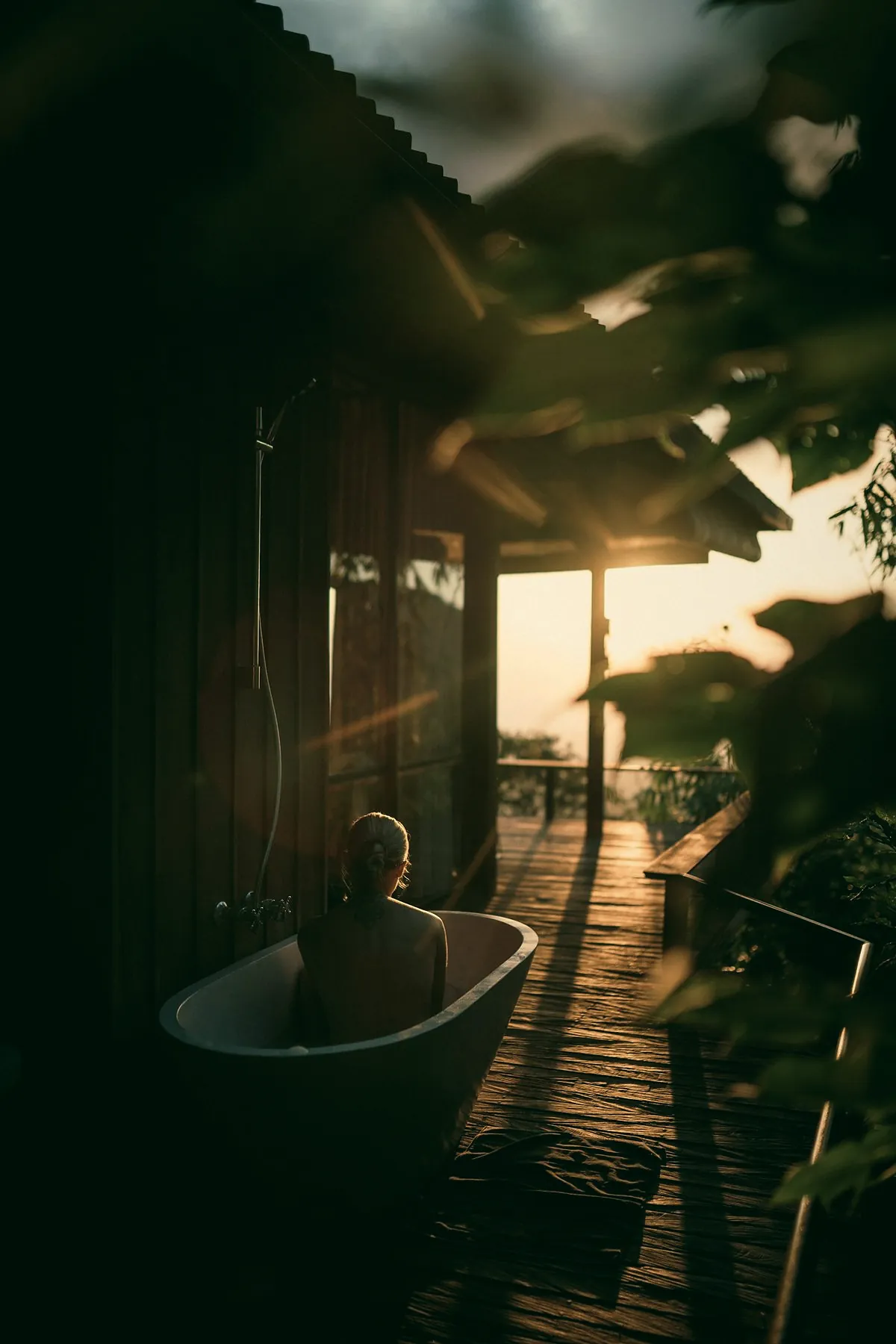 A person in a white bathtub on a dark wooden terrace, with the low sun behind the trees