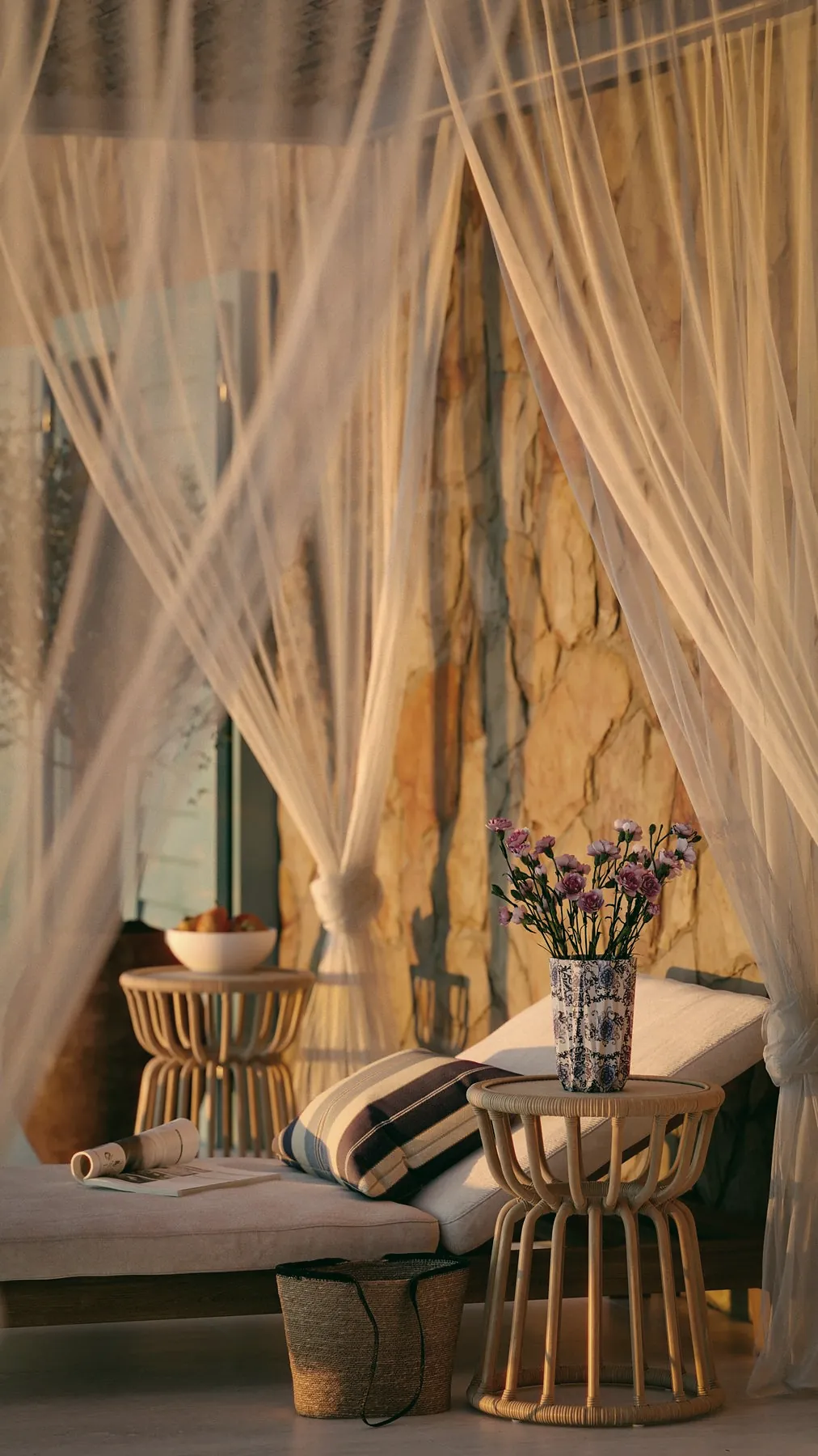 A daybed with cushions behind sheer curtains, a rattan table and a vase of flowers