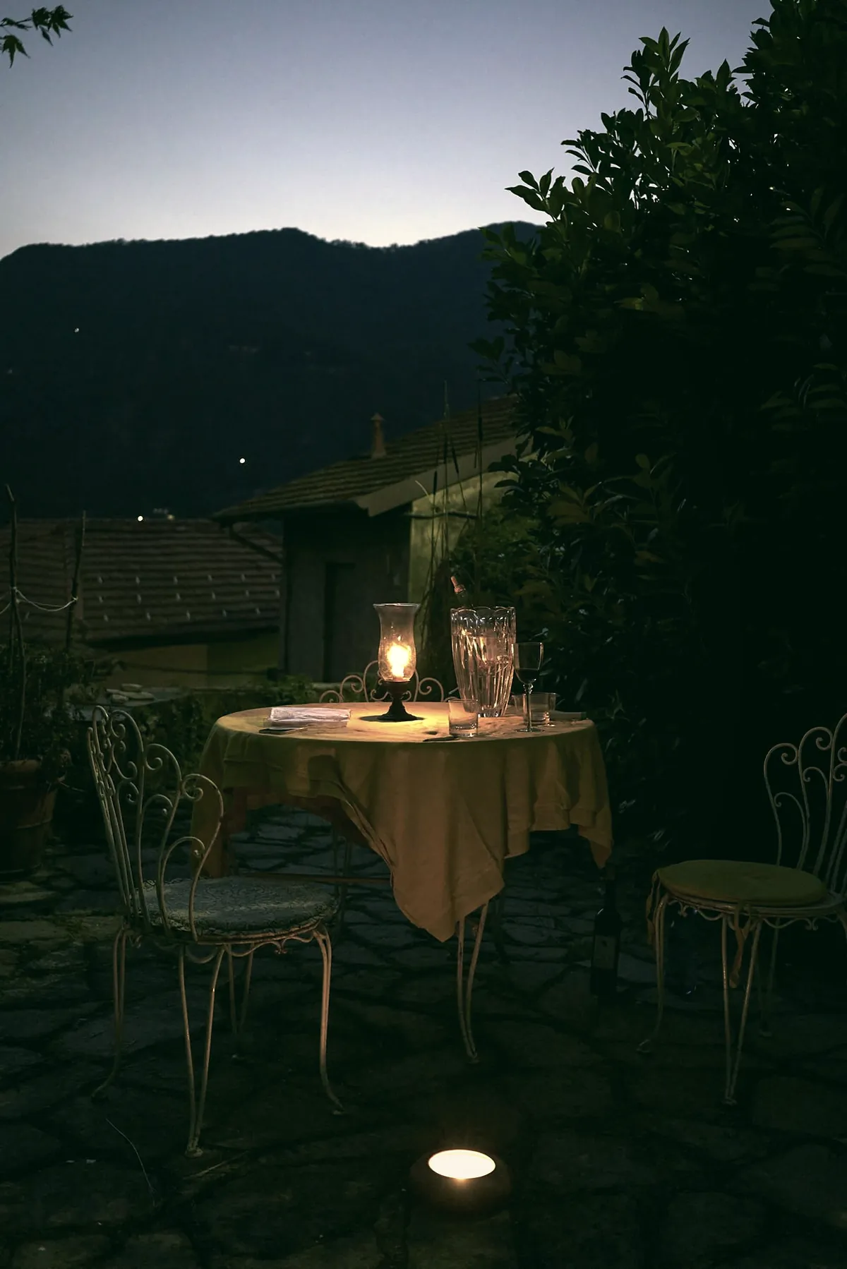 A table for two with a candle lamp on a stone terrace at dusk, with dark hills behind