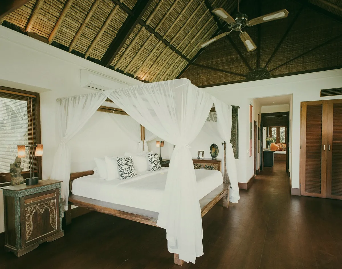 A bedroom with a canopy bed under a tall alang-alang roof, with wooden floors and white nets