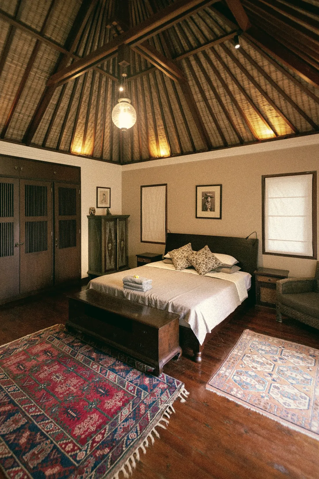A bedroom with a high wooden roof, a pendant lamp, a wooden bed and red rugs on a timber floor