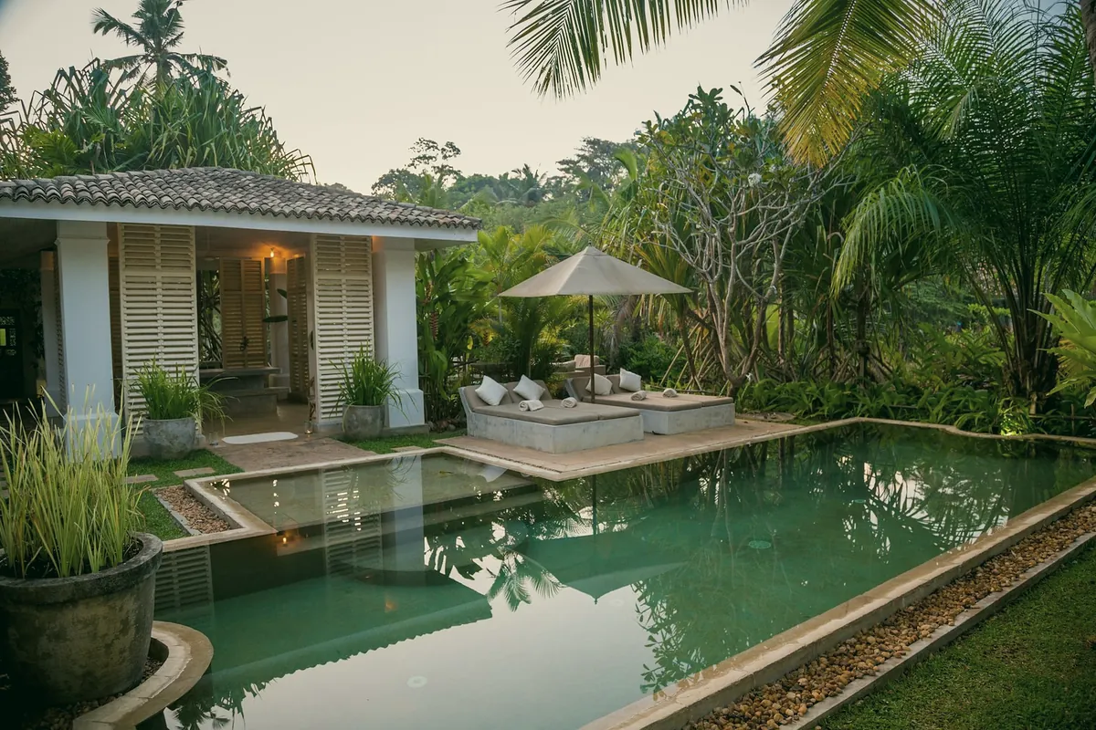 A long green pool in a tropical garden, with a white pavilion and two loungers under an umbrella