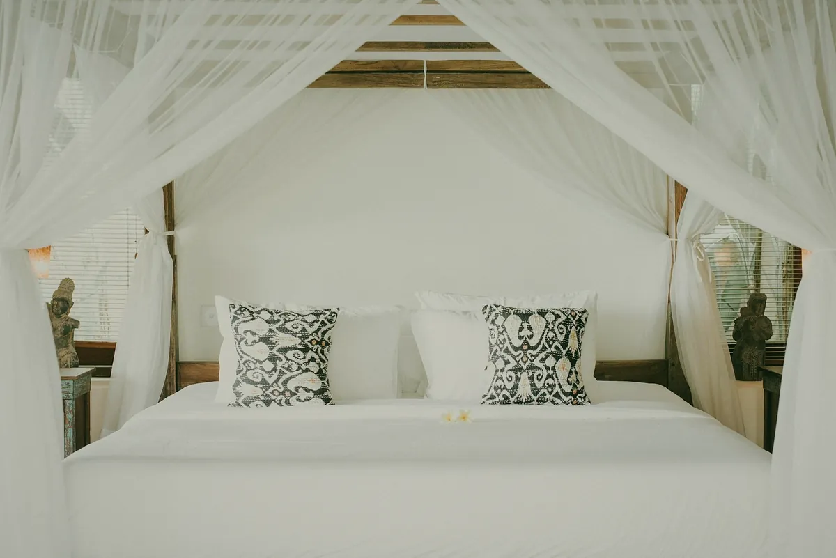 A white bed under a wooden canopy with white curtains and two patterned cushions
