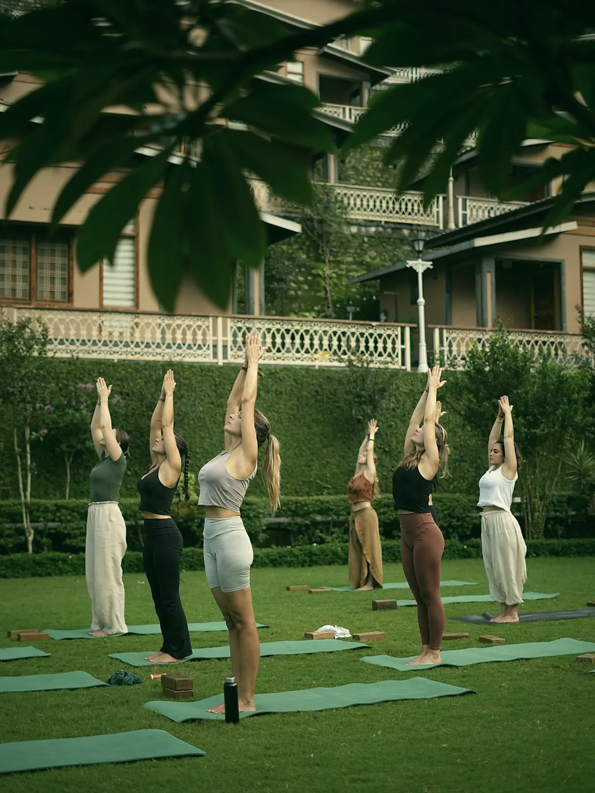 Six women standing on mats on a lawn with their arms raised, in front of a garden and a house