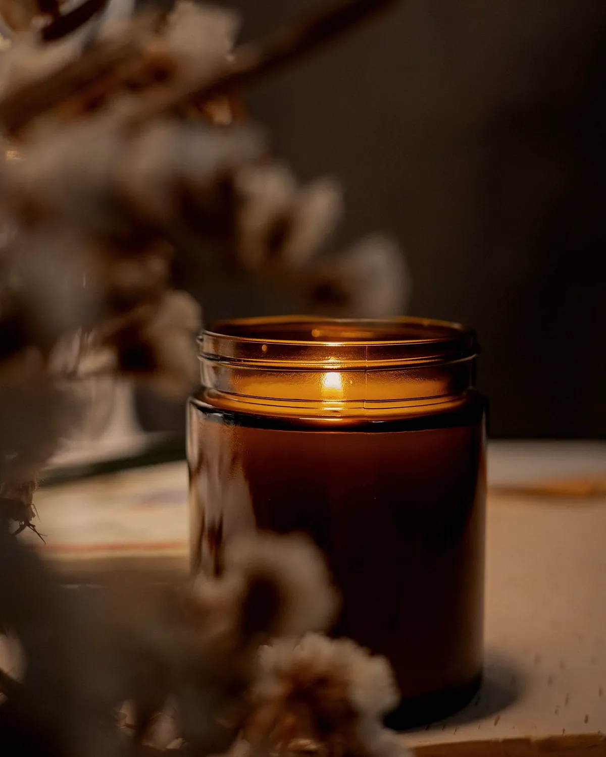 A lit candle in an amber jar behind a spray of dried flowers in the evening