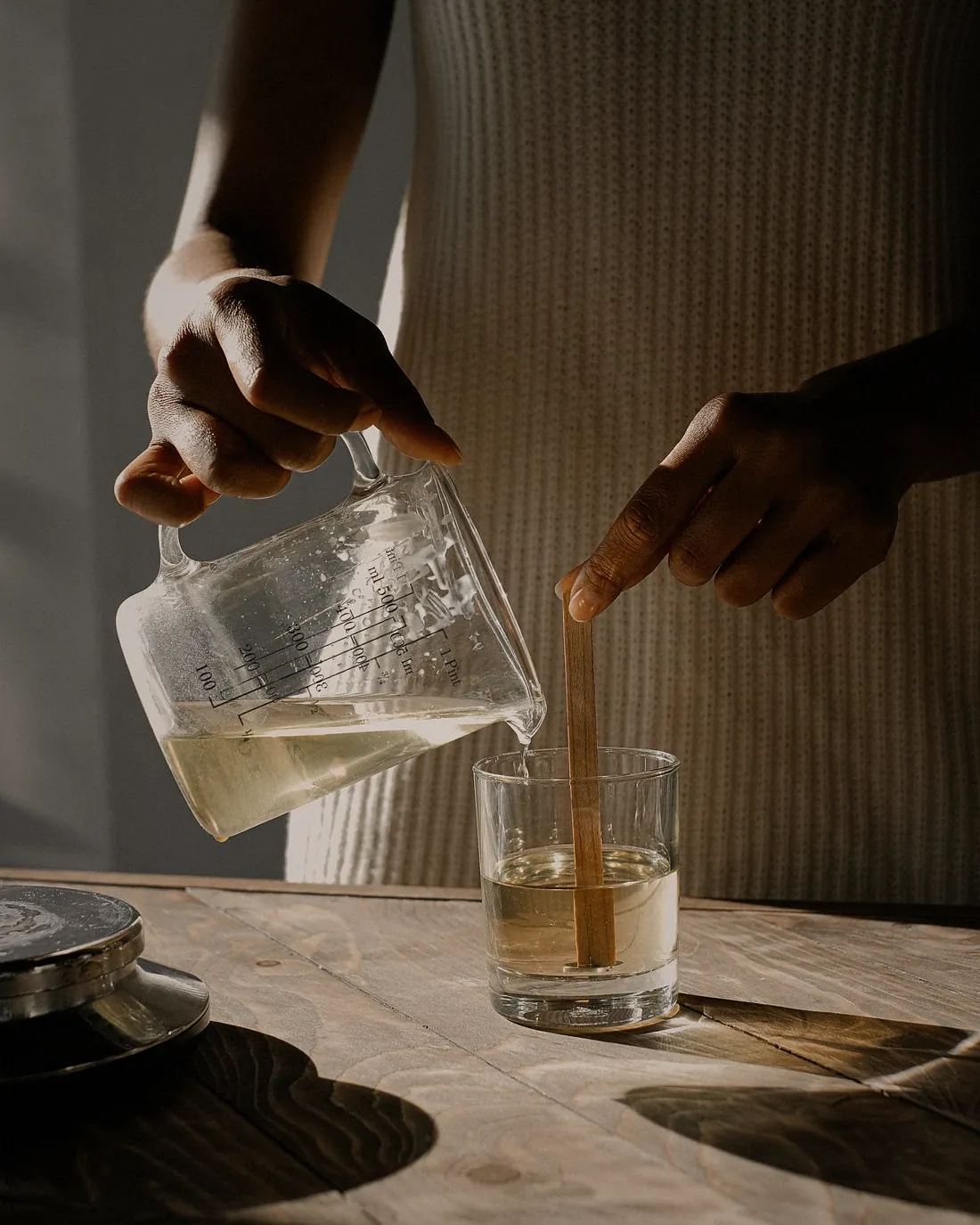 Hands pouring melted wax from a glass jug into a jar on a wooden bench